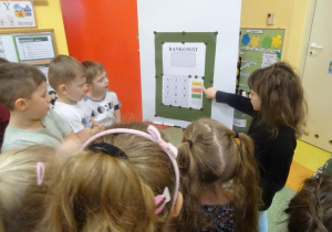 Grupa dzieci stoi wokół tablicy na której przywieszony jest szablon bankomatu.